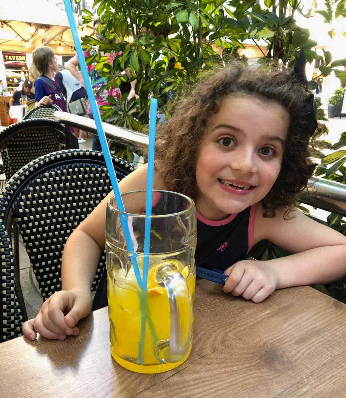girl with dark curly hair smiling for the camera - she has a yellow drink in a large glass with blue straws in front of her
